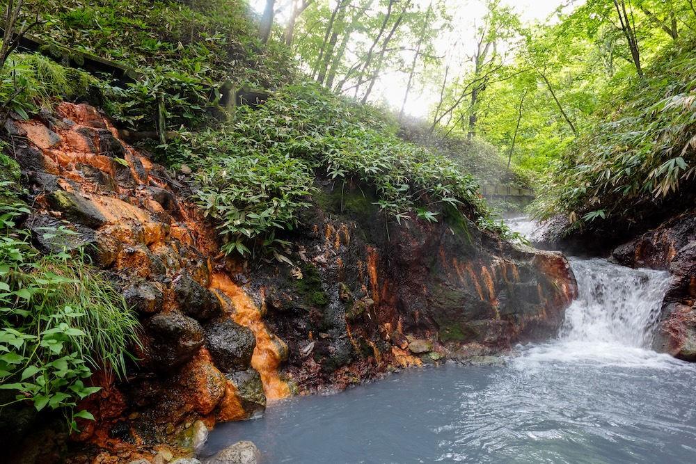 Orange Mineral-Stained Rocks and Cascade at the Oyunuma River Natural Footbath, Noboribetsu Onsen, Hokkaido, Japan Orange mineral-stained rocks and bamboo grass frame a small cascade tumbling into a milky blue hot-spring pool under the forest at the Oyunuma River Natural Footbath in Noboribetsu Onsen, Hokkaido, Japan.