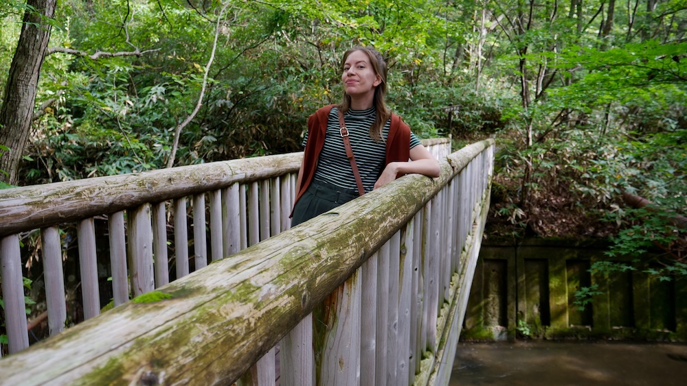 Puente de madera en el bosque cerca del baño de pies del río Oyunuma, Noboribetsu Onsen, Japón – Audrey Bergner Audrey Bergner apoyada en la baranda de troncos cubiertos de musgo de un puente peatonal de madera en medio del bosque verde cerca del baño de pies natural del río Oyunuma, con un arroyo y un muro de piedra abajo, en Noboribetsu Onsen, Hokkaido, Japón.