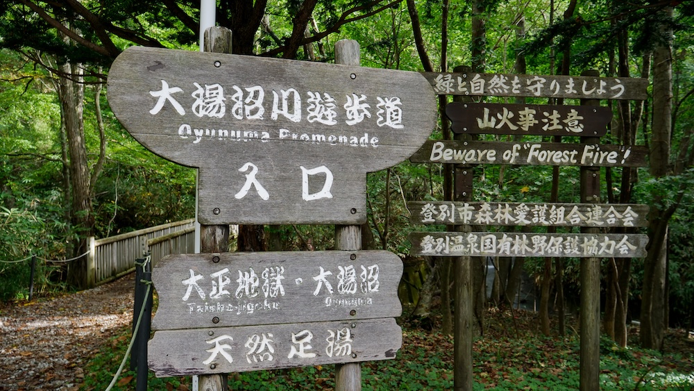 Cartel de entrada al paseo del río Oyunuma hacia Taisho Jigoku y baño de pies, Noboribetsu Onsen, Japón Carteles de madera marcan la entrada del paseo del río Oyunuma y señalan hacia Taisho Jigoku, Oyunuma y el baño de pies natural, junto a un sendero frondoso y un puente peatonal en Noboribetsu Onsen, Hokkaido, Japón.