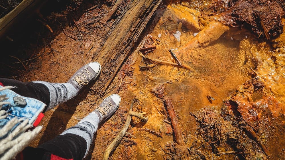 Audrey Bergner beside an ochre mineral spring at the Paint Pots, Kootenay National Park, British Columbia, Canada Audrey Bergner’s feet beside a shallow ochre-coloured mineral spring at the Paint Pots in Kootenay National Park, British Columbia, Canada, with orange-stained earth, clear water, roots and weathered wood visible.