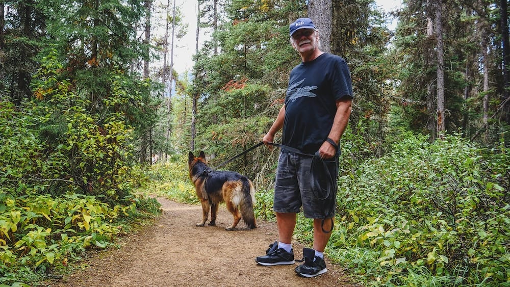 Daniel Bergner and Togo on the Paint Pots Trail in Kootenay National Park, British Columbia, Canada Daniel Bergner stands with Togo on the forested Paint Pots Trail in Kootenay National Park, British Columbia, Canada, with a narrow dirt path, dense conifers and green undergrowth surrounding them.