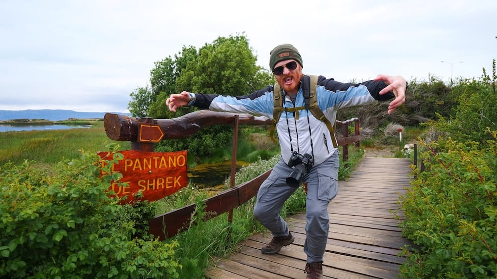 Pantano de Shrek Wetland Boardwalk in El Calafate, Patagonia, Argentina, with Samuel Jeffery Samuel Jeffery poses playfully beside the wooden Pantano de Shrek sign on a wetland boardwalk in El Calafate, Santa Cruz Province, Patagonia, Argentina, wearing hiking clothes and carrying a camera.