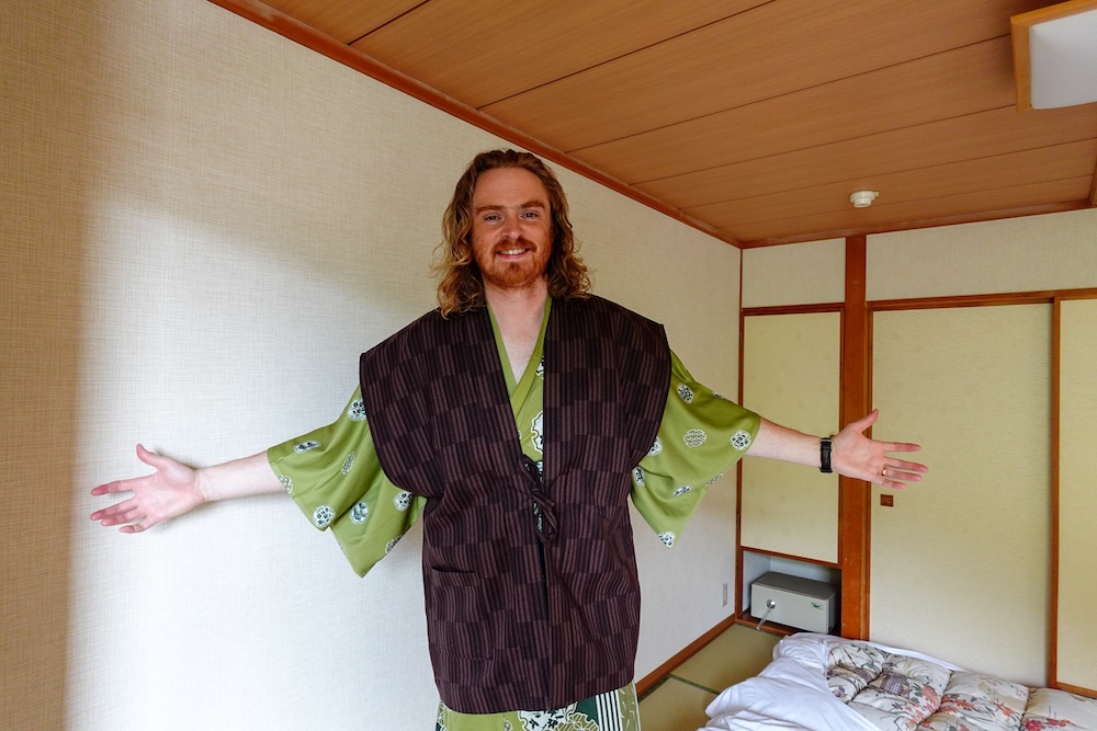 Samuel Jeffery spreads his arms wide in a green yukata and dark vest inside a tatami-floored Japanese-style room with sliding doors and a laid-out futon at Park Hotel Miyabitei, Noboribetsu Onsen, Hokkaido, Japan.