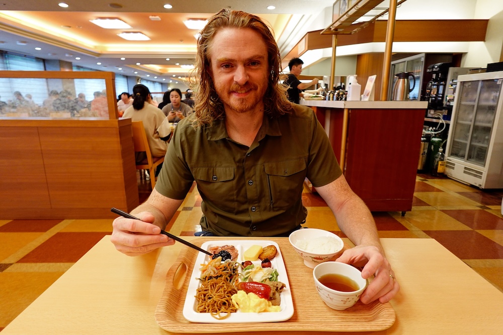 Samuel Jeffery disfrutando del buffet de la cena en Park Hotel Miyabitei, Noboribetsu Onsen, Hokkaido, Japón Samuel Jeffery sentado en el comedor buffet de Park Hotel Miyabitei, Noboribetsu Onsen, Hokkaido, Japón, con palitos en la mano frente a una bandeja con fideos, arroz, verduras, platos calientes variados y té.