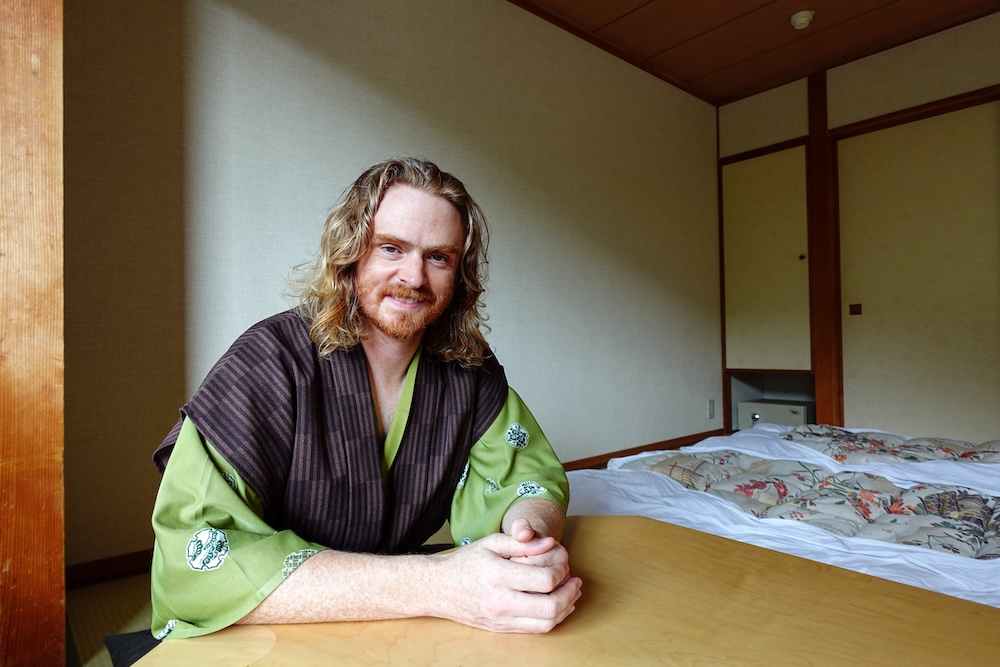 Samuel Jeffery con un yukata a rayas oscuras y mangas verdes, sentado ante una mesa de madera dentro de una habitación de estilo japonés del Park Hotel Miyabitei, en Noboribetsu Onsen, Hokkaido, Japón, con futones preparados detrás de él.