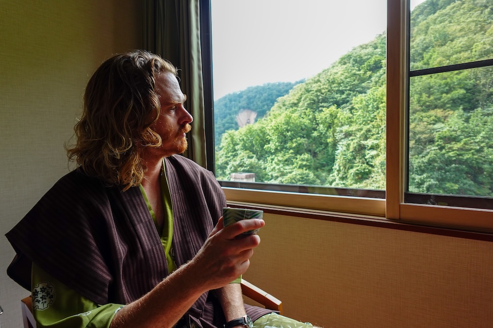 Samuel Jeffery tomando té en una habitación de estilo japonés de Park Hotel Miyabitei, Noboribetsu Onsen, Hokkaido, Japón Samuel Jeffery vestido con una bata de estilo japonés y sosteniendo una taza de té junto a la ventana de su habitación en Park Hotel Miyabitei, Noboribetsu Onsen, Hokkaido, Japón, con vistas a colinas cubiertas de árboles verdes.