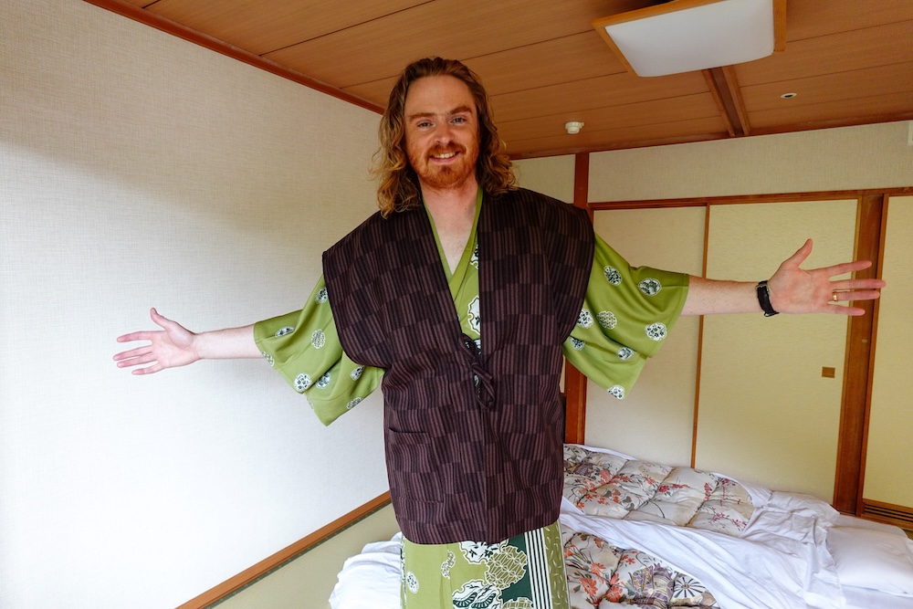 Samuel Jeffery Wearing Yukata at Park Hotel Miyabitei Traditional Japanese Room in Noboribetsu Onsen, Hokkaido Samuel Jeffery smiles with his arms outstretched, wearing a green patterned yukata and dark vest inside a traditional Japanese-style room with futon bedding at Park Hotel Miyabitei in Noboribetsu Onsen, Hokkaido, Japan.