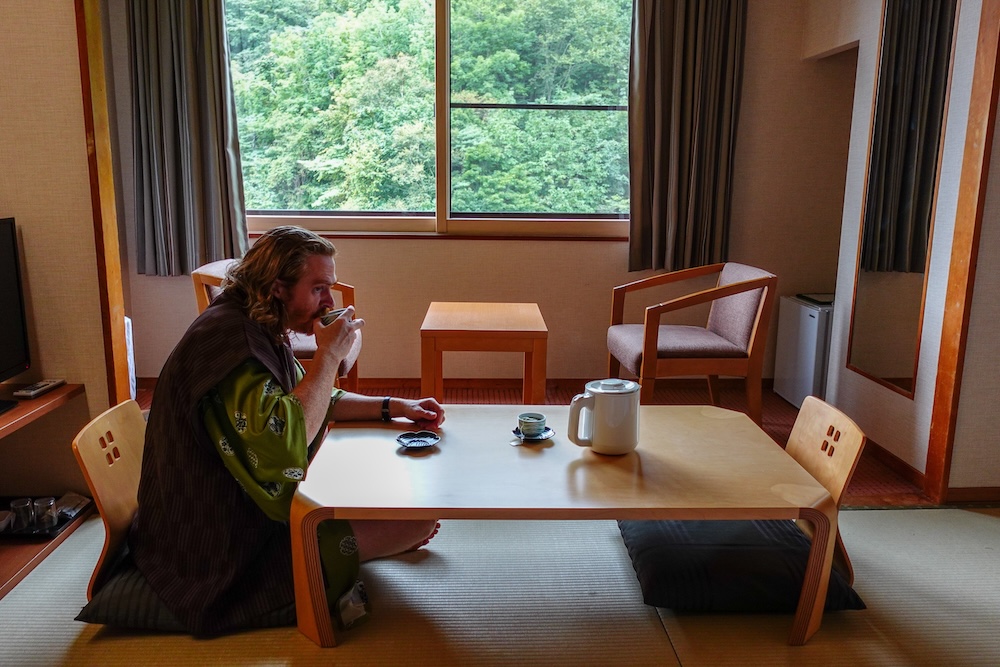 Samuel Jeffery vestido con un yukata tradicional y tomando té japonés en una mesa baja de madera dentro de una habitación con tatami del Park Hotel Miyabitei, en Noboribetsu Onsen, Hokkaido, Japón, con asientos en el piso y vistas al bosque por la ventana.