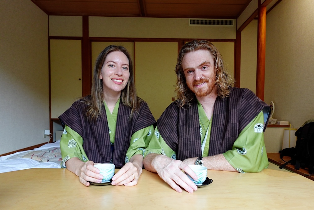 Samuel Jeffery y Audrey Bergner tomando té con yukata en el Park Hotel Miyabitei, Noboribetsu Onsen, Hokkaido, Japón Audrey Bergner y Samuel Jeffery vestidos con yukata verdes y chaquetas oscuras a rayas mientras toman té japonés frente a una mesa baja de madera en su habitación tradicional del Park Hotel Miyabitei, Noboribetsu Onsen, Hokkaido, Japón.