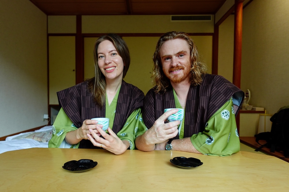 Samuel Jeffery and Audrey Bergner Enjoying Tea in Yukata at Park Hotel Miyabitei, Noboribetsu Onsen, Hokkaido, Japan Samuel Jeffery and Audrey Bergner smile while holding cups of Japanese tea at a low wooden table, wearing green patterned yukata and dark vests inside their traditional Japanese-style room at Park Hotel Miyabitei in Noboribetsu Onsen, Hokkaido, Japan.