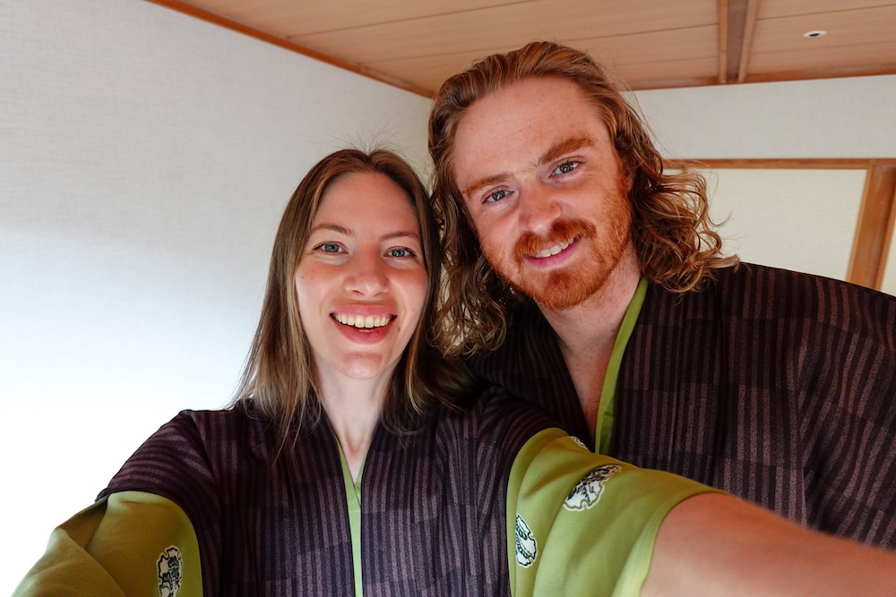 Samuel Jeffery y Audrey Bergner sonriendo para una selfie en su habitación de estilo japonés del Park Hotel Miyabitei, en Noboribetsu Onsen, Hokkaido, Japón, vestidos con yukata a rayas oscuras, mangas verdes y cuellos del mismo color.