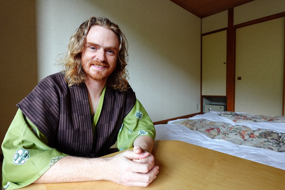 Futons laid out in a tatami room at Park Hotel Miyabitei, Noboribetsu Onsen, Hokkaido, Japan – Samuel Jeffery Samuel Jeffery in a green yukata and dark vest sits at a low wooden table in a tatami room. Futons with floral duvets are laid out behind him at Park Hotel Miyabitei, Noboribetsu Onsen, Hokkaido, Japan.