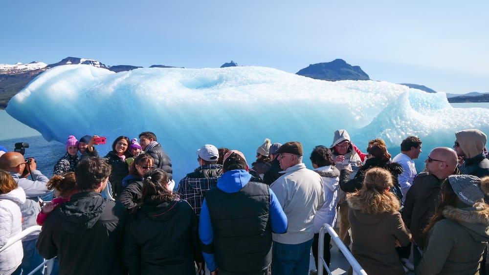 Cubierta repleta en un crucero a los glaciares por el Lago Argentino cerca de El Calafate, Santa Cruz, Argentina Una cubierta de barco repleta de pasajeros abrigados con gorros y camperas fotografiando un enorme témpano azul y blanco flotando en el Lago Argentino, con picos nevados a lo lejos, en un crucero a los glaciares cerca de El Calafate, Santa Cruz, Patagonia argentina.