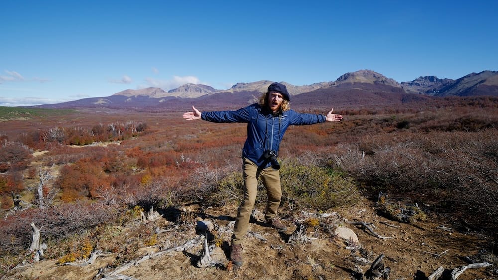 Samuel Jeffery está con los brazos abiertos sobre un valle patagónico de colores otoñales cerca de Esquel, Chubut, Argentina, con matorral bajo, cordones montañosos distantes y un cielo azul despejado.