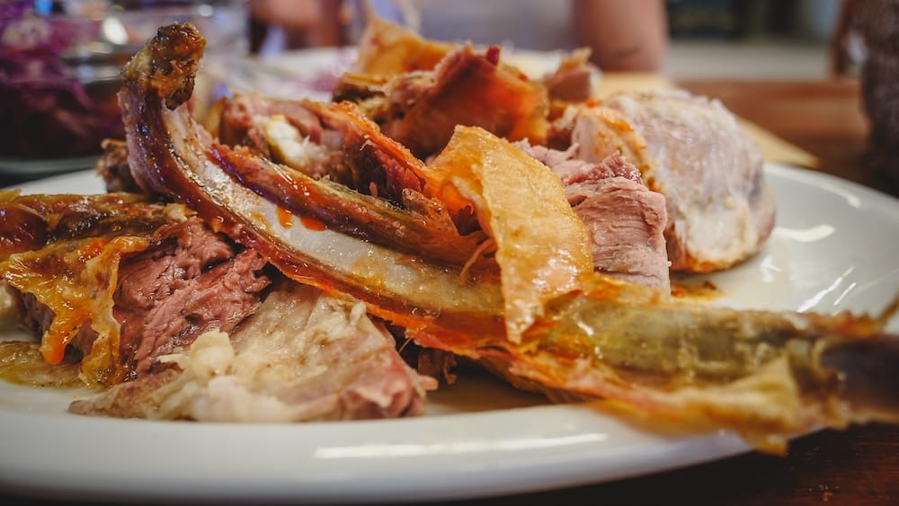 Close-up of roasted Patagonian lamb served on a white platter during a Península Valdés wildlife tour in Chubut, Patagonia, Argentina, with crisp skin, ribs and tender carved meat filling the frame.
