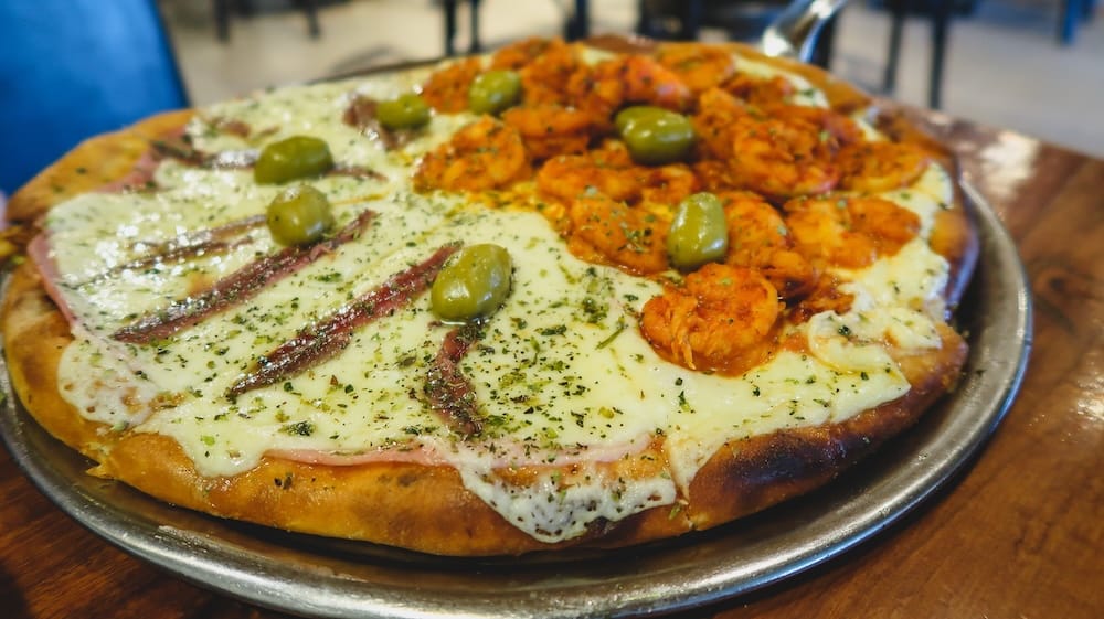Patagonian seafood and anchovy pizza in Puerto Madryn with shrimp, olives, garlic, and thin crust baked in coastal Argentina Half-and-half Patagonian seafood and anchovy pizza in Puerto Madryn featuring shrimp, anchovies, green olives, melted mozzarella, garlic, and herbs on a thin crust served on a metal tray along Argentina’s Atlantic coast.