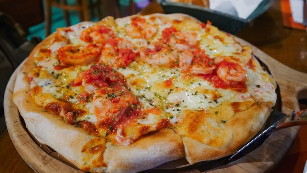 Shrimp pizza in Comodoro Rivadavia Patagonia with mozzarella tomato sauce and thick crust served on wooden board Patagonian shrimp pizza served in Comodoro Rivadavia Argentina with melted mozzarella, tomato sauce, herbs, and whole shrimp on a thick crust presented on a wooden board in a casual coastal restaurant setting.