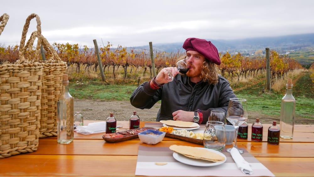 Samuel Jeffery, wearing a burgundy boina, tastes red wine at an outdoor picnic table among the vines at Patagonian Wines in El Hoyo, Chubut, Argentine Patagonia, with cheese and cured meats laid out.