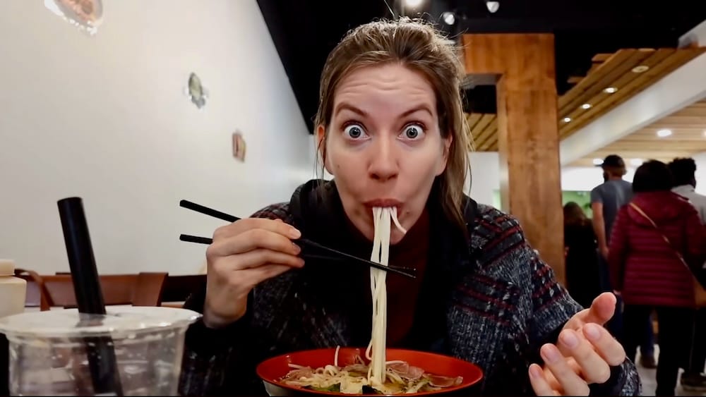 Audrey Bergner makes an exaggerated wide-eyed face while slurping pho noodles with chopsticks at Peak Cafe in Banff, Alberta, Canada, with a bubble tea visible in the foreground.
