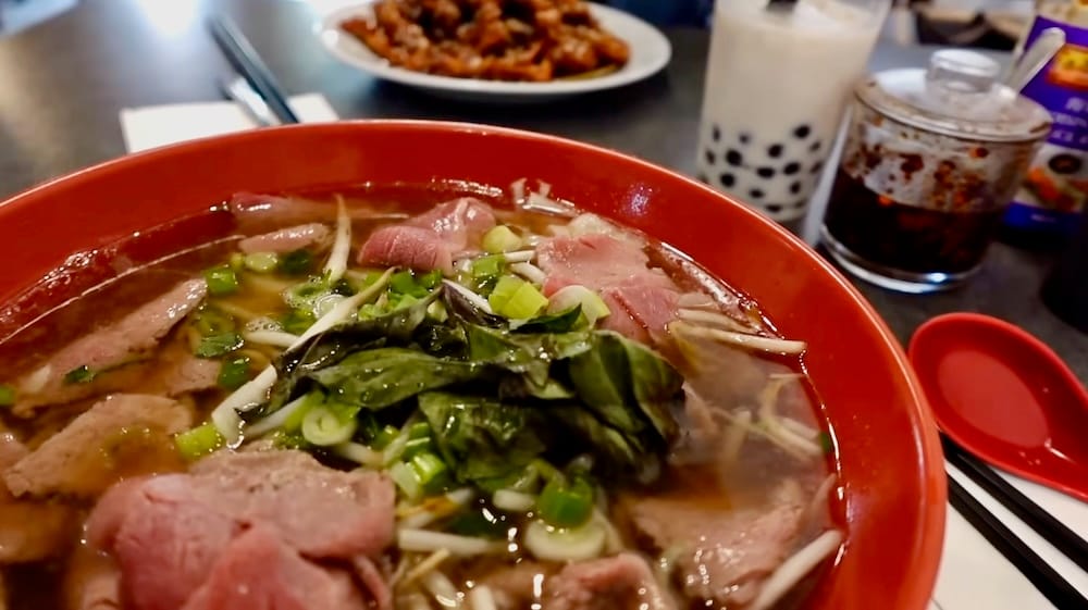 A table spread at Peak Cafe in Banff, Alberta, Canada, showing a bowl of beef pho in the foreground with ginger beef and a bubble tea visible in the background.