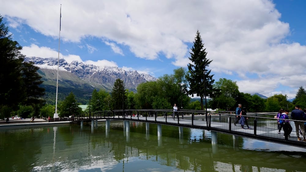 Pedestrian bridge crossing a reflective pond in El Bolsón, Argentina, with visitors walking across it and snow-capped mountain peaks rising behind the trees under a partly cloudy sky.