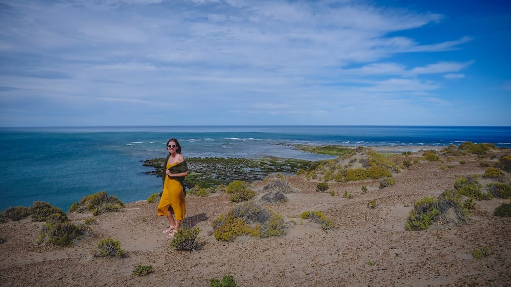 Audrey Bergner stands at a sandy coastal steppe viewpoint in Península Valdés, Chubut, Patagonia, Argentina, with low scrub, rocky tidal flats and the broad blue Atlantic Ocean stretching behind her.