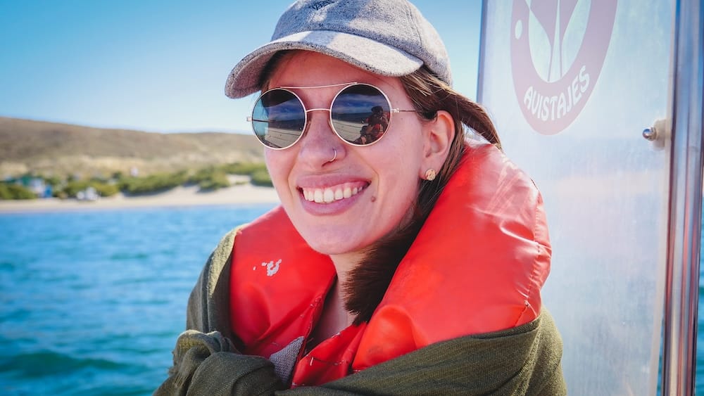 Audrey Bergner a bordo de una embarcación de fauna en Península Valdés, Chubut, Patagonia, Argentina Audrey Bergner sonríe a bordo de una embarcación para observar fauna en Península Valdés, Chubut, Patagonia, Argentina, con chaleco salvavidas rojo, gorra gris y anteojos de sol, frente al agua azul y la costa baja.