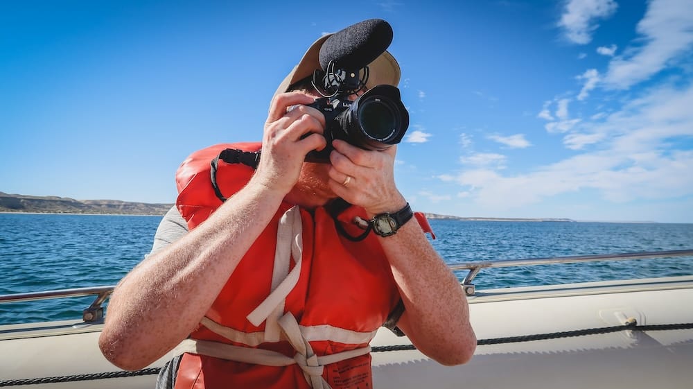 Samuel Jeffery fotografiando fauna desde un barco en Península Valdés, Chubut, Patagonia, Argentina Samuel Jeffery fotografía fauna desde una embarcación pequeña en Península Valdés, Chubut, Patagonia, Argentina, con chaleco salvavidas rojo, el mar abierto y acantilados costeros bajos detrás.