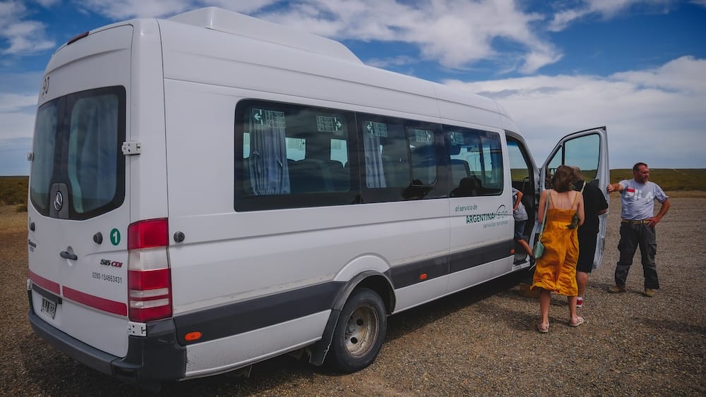 Audrey Bergner boards a white wildlife tour van on the open steppe of Península Valdés, Chubut, Patagonia, Argentina, while other passengers gather beneath a bright blue and cloud-streaked sky.