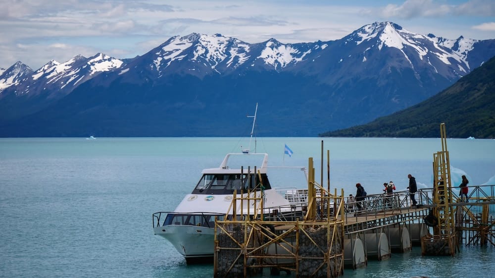 Glacier boat departing Perito Moreno in Santa Cruz, Patagonia, beneath snowcapped mountain peaks A glacier tour boat departs from the dock at Perito Moreno Glacier in Santa Cruz, Patagonia, Argentina, with passengers boarding beneath jagged snowcapped mountains and pale blue lake water.