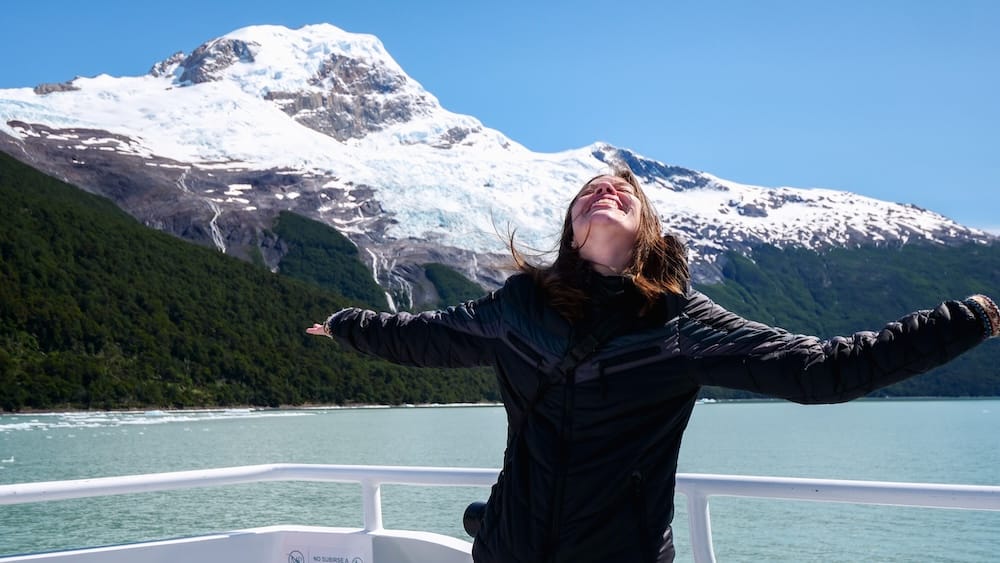 Perito Moreno Glacier Boat Tour Near El Calafate, Patagonia, Argentina, with Audrey Bergner Audrey Bergner stands with her arms open on a glacier tour boat near El Calafate, Santa Cruz, Patagonia, Argentina, with turquoise water, forested slopes and a snow-covered glacier beneath a clear blue sky.