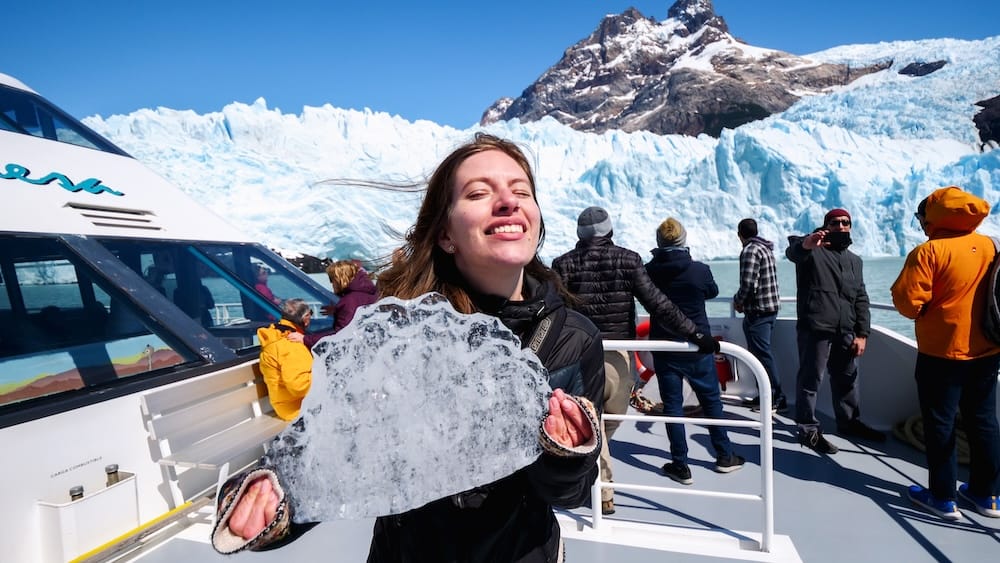 Audrey Bergner holds a large piece of glacial ice on the deck of a boat beside Perito Moreno Glacier in Santa Cruz, Patagonia, Argentina, with passengers and towering blue ice behind her.