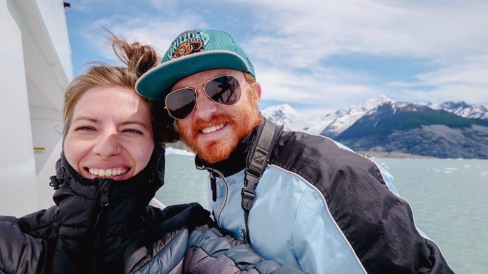 Samuel Jeffery and Audrey Bergner smile aboard a glacier boat near Perito Moreno in Santa Cruz, Patagonia, Argentina, with pale glacial water and snow-covered mountain peaks behind them.