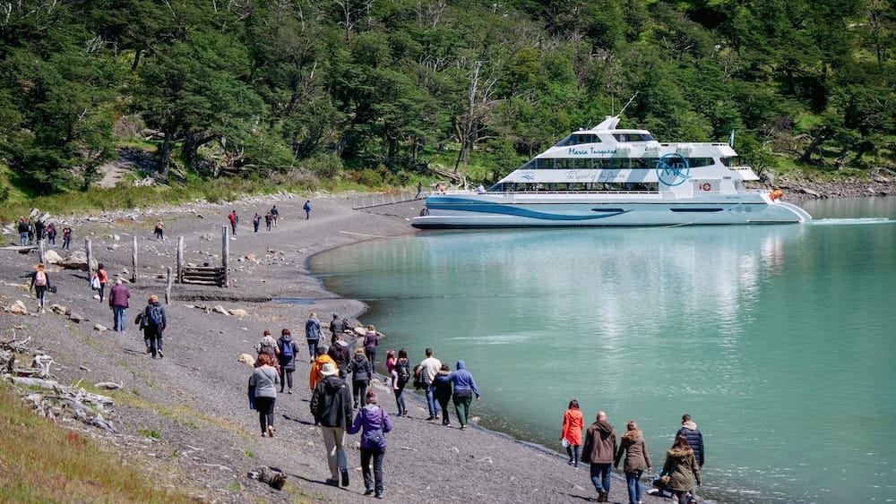 Tourists walk along a rocky lakeshore to board a white catamaran near Perito Moreno Glacier in Santa Cruz, Patagonia, Argentina, beside pale turquoise glacial water and dense forest.
