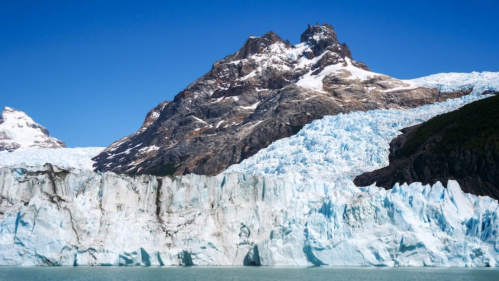 Towering ice wall and rugged peaks at Perito Moreno Glacier in Patagonia, Argentina Towering blue-white ice walls of Perito Moreno Glacier rise above Lago Argentino in Santa Cruz, Patagonia, Argentina, beneath rugged snow-dusted mountain peaks and a clear blue sky.