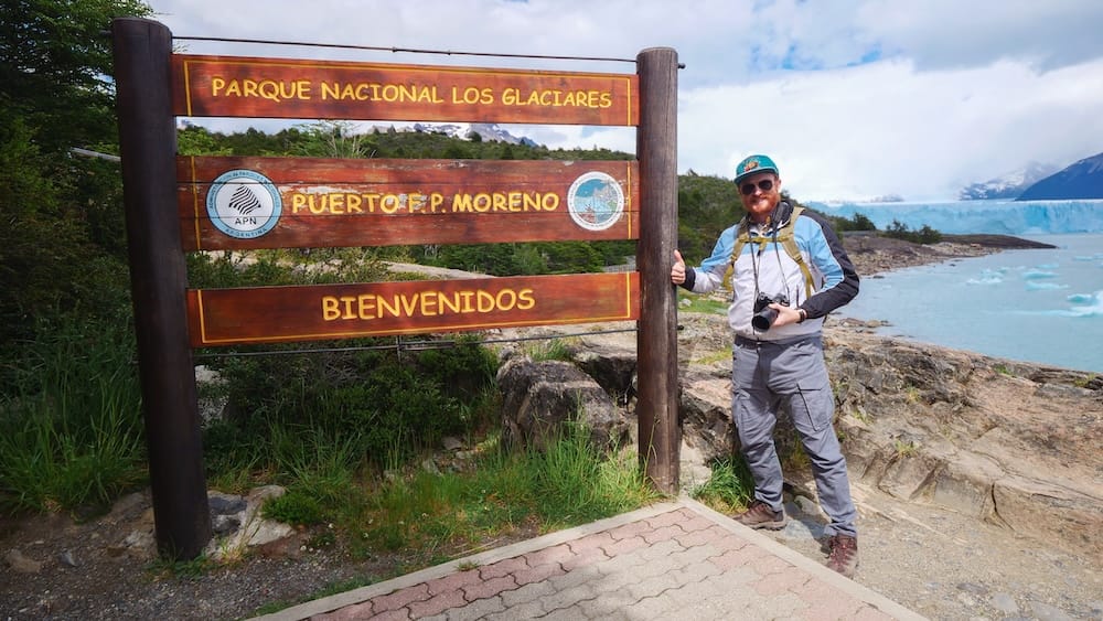 Samuel Jeffery at the Perito Moreno Glacier Entrance Sign, Los Glaciares National Park, Santa Cruz Samuel Jeffery gives a thumbs-up beside the "Parque Nacional Los Glaciares, Puerto F.P. Moreno" welcome sign, with Perito Moreno Glacier and floating ice visible on Lake Argentino behind him in Santa Cruz, Argentine Patagonia.