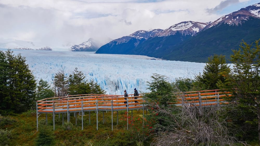 Two travelers walk along elevated wooden boardwalks overlooking Perito Moreno Glacier in Los Glaciares National Park, Santa Cruz, Patagonia, Argentina, with blue ice, forest and snow-streaked mountains behind them.