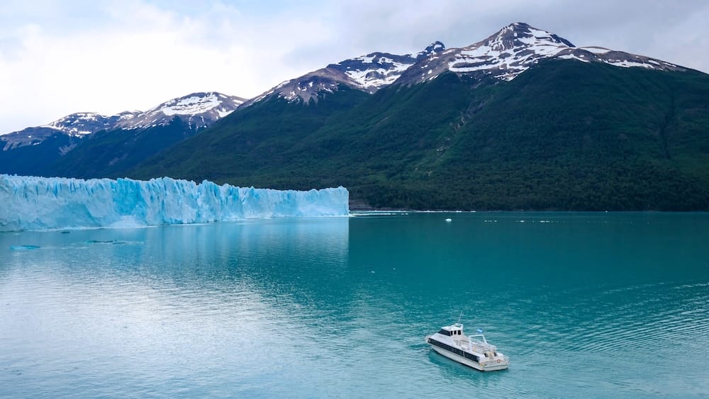 A sightseeing boat floats on turquoise water near the blue ice wall of Perito Moreno Glacier in Los Glaciares National Park, Santa Cruz, Patagonia, Argentina, with forested mountains rising behind.