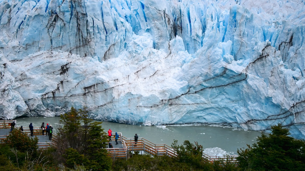 Perito Moreno Glacier towering above viewing boardwalks in El Calafate, Patagonia, Argentina, with massive blue ice walls calving into a glacial lake and visitors standing at the official viewpoint in Los Glaciares National Park.