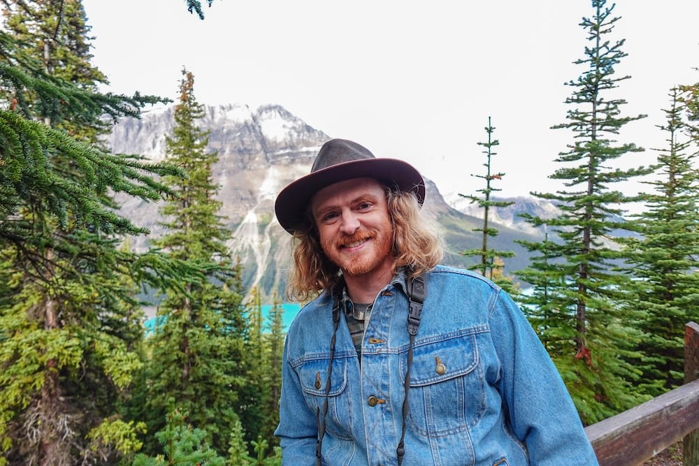 Samuel Jeffery está en un mirador boscoso sobre Peyto Lake, junto a la Icefields Parkway en Alberta, Canadá, con agua turquesa entre los árboles y laderas claras detrás.