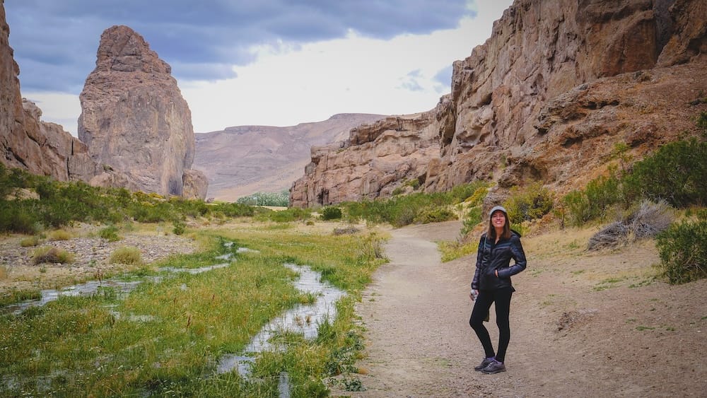 Audrey Bergner stands on a dirt trail at Piedra Parada in Chubut, Patagonia, Argentina, with a towering volcanic monolith, rocky canyon walls and marshy green flats stretching through the dramatic arid landscape.