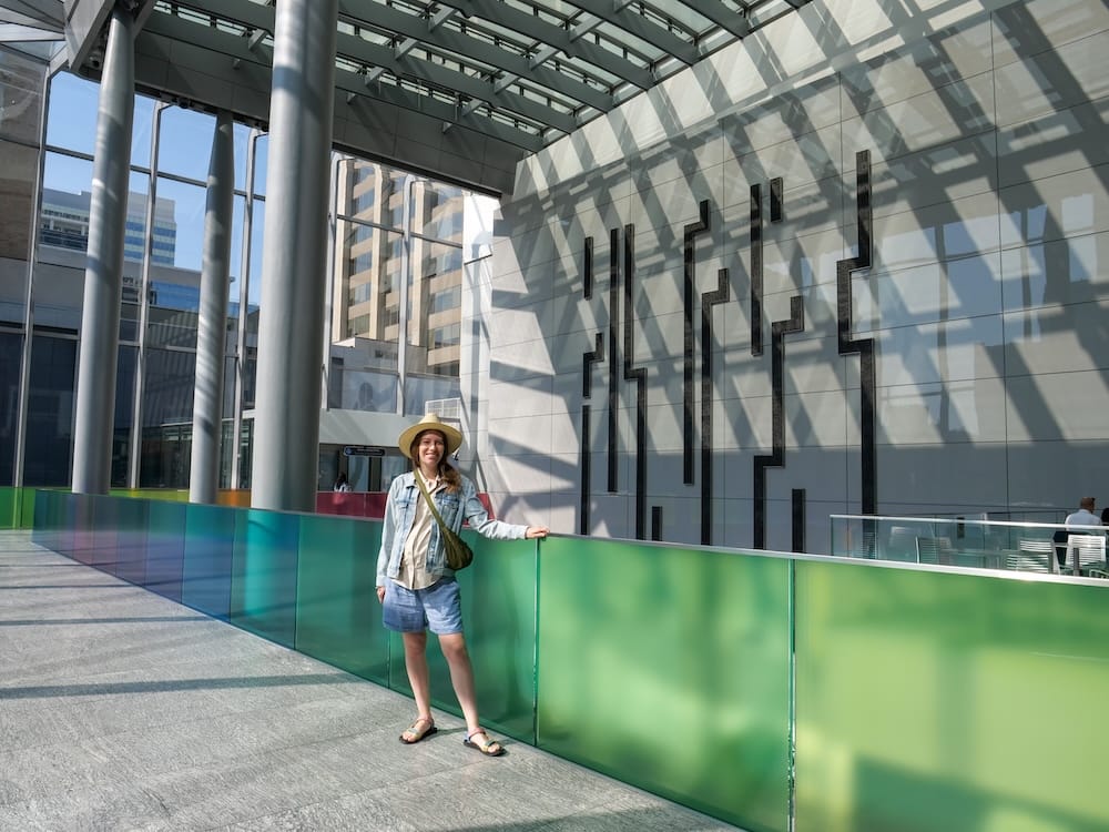 Calgary +15 Skywalk Alberta Canada Indoor Pedestrian Network with Audrey Bergner Downtown Audrey Bergner stands inside Calgary’s +15 Skywalk in Downtown Calgary, Alberta, Canada, beside colourful glass railings, tall columns and geometric wall art beneath a glass roof.