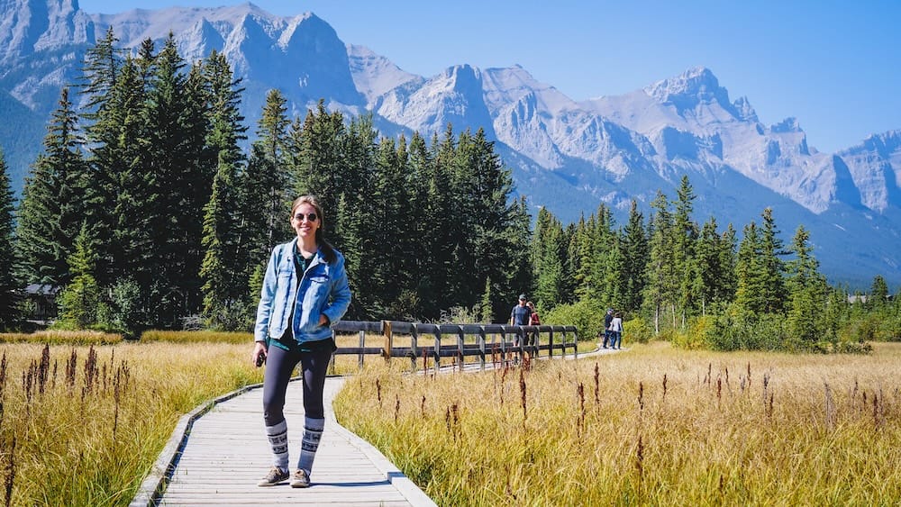 Audrey Bergner walks along Policeman’s Creek Boardwalk in Canmore, Alberta, Canada, with golden wetland grasses, evergreen forest and rugged Canadian Rockies peaks rising behind her.