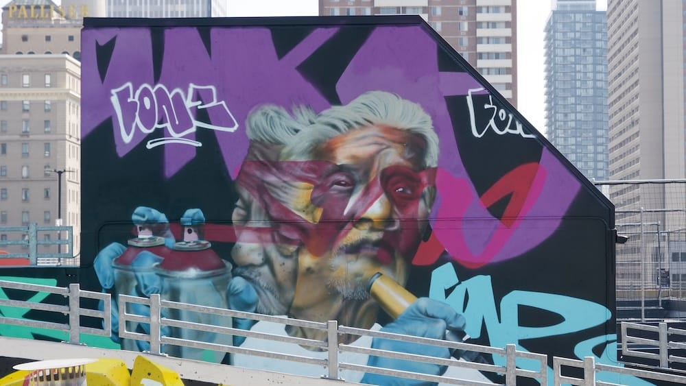 Un gran mural del retrato de un hombre mayor con marcas pintadas en la cara sosteniendo latas de pintura en aerosol, sobre un fondo negro y violeta, en una pared de la terraza en High Park en Calgary, Alberta, Canadá, con el hotel Fairmont Palliser visible a lo lejos.