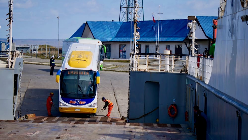 Bus Sur Coach to Ushuaia Boarding Ferry at Primera Angostura, Strait of Magellan, Magallanes, Chilean Patagonia A white and yellow Bus Sur double-decker coach marked "USHUAIA" boards a car ferry crossing the Strait of Magellan near Primera Angostura in Chile's Magallanes Region, guided by dock crew in orange overalls under bright midday sun.