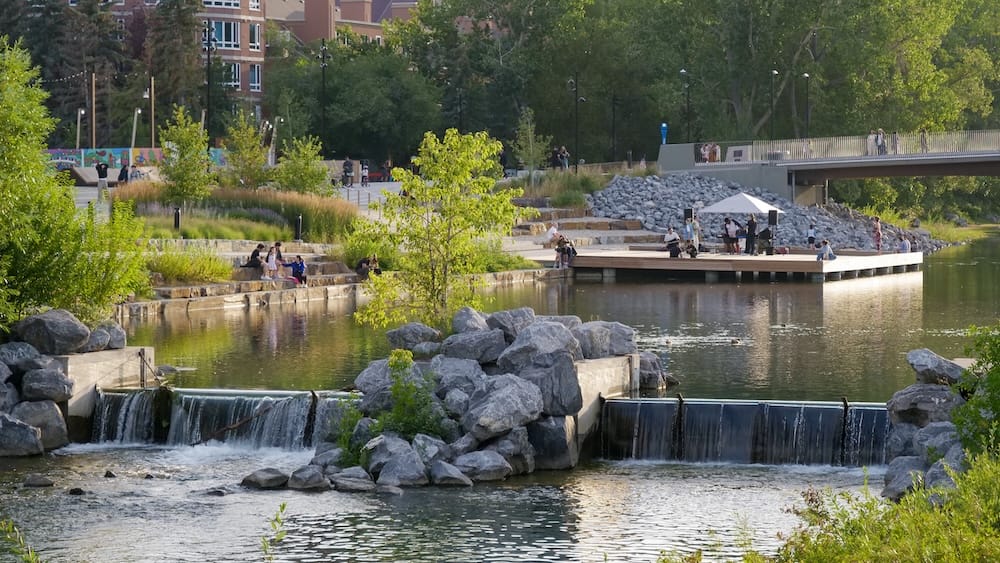 Prince’s Island Park in Calgary, Alberta, Canada, with landscaped riverfront paths, small waterfalls, rocky banks, trees and people relaxing beside the water on a sunny day.