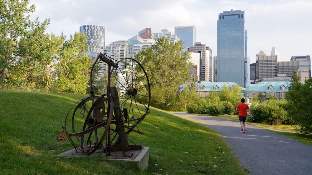 Sendero de Prince’s Island Park y skyline del centro de Calgary, Alberta, Canadá Sendero para caminar en Prince’s Island Park, Calgary, Alberta, Canadá, con césped verde, árboles, una escultura metálica y el skyline del centro elevándose detrás del parque junto al río.