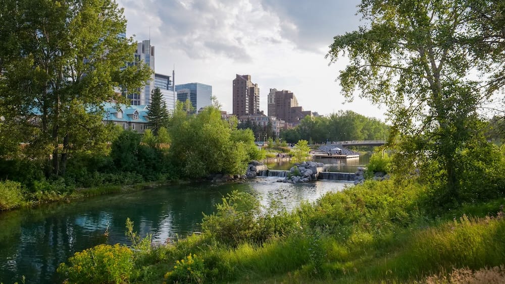 Prince's Island Park River Lagoon and Downtown Skyline, Calgary, Alberta, Canada Tranquil lagoon and stone weir in Prince's Island Park framed by green trees and grass, with downtown Calgary high-rises visible under a cloud-dappled sky.