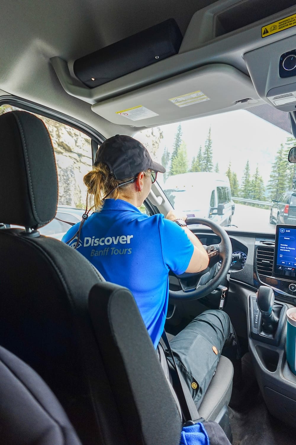 A private Banff tour guide driving through the Canadian Rockies in Alberta, Canada, with forested mountain roads visible ahead from inside the tour vehicle.