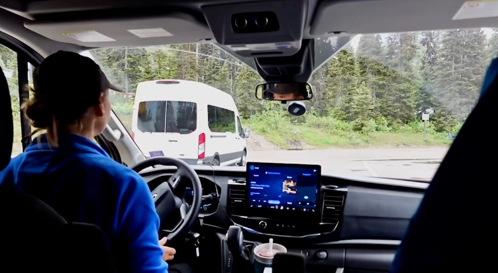 View from inside a private tour van as it drives into the Lake Louise parking area in Banff National Park, Alberta, Canada, with another tour shuttle visible ahead on the access road.